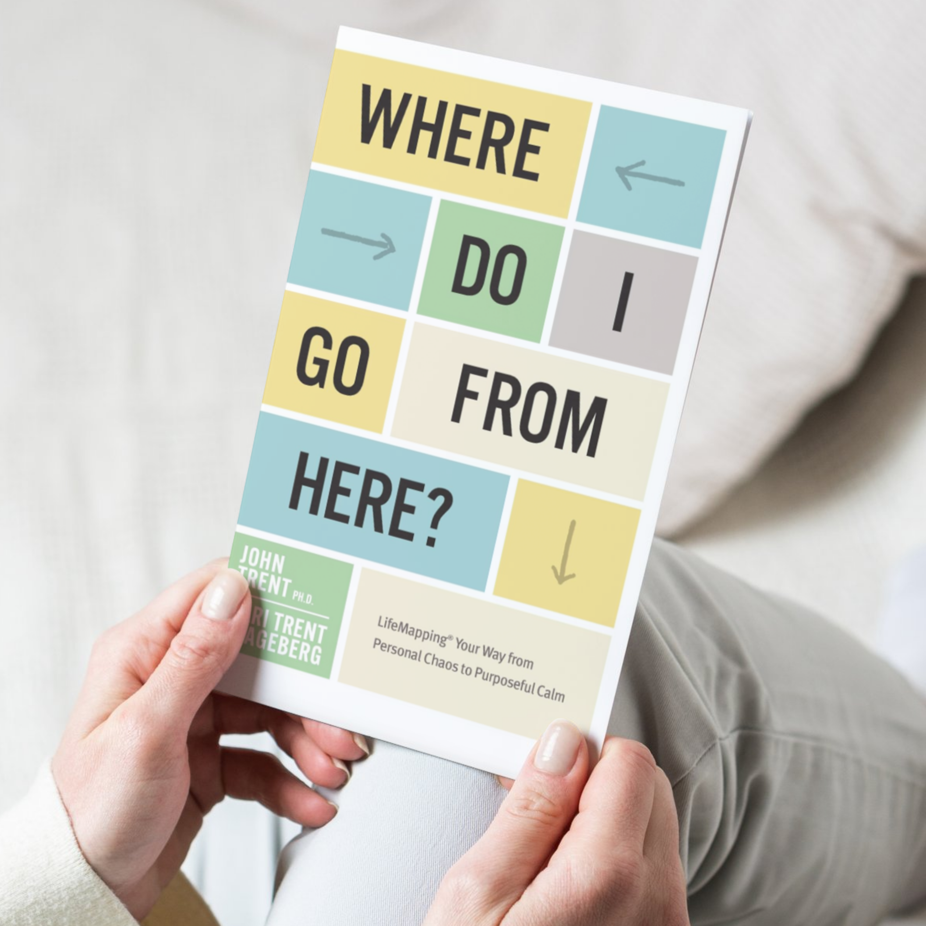 hands with a white shirt and grey pants holding the book cover for 'Where Do I Go From Here?' by John Trent and Kari Trent Stageberg, featuring a collage design with various text and arrows on a colorful background.