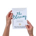Hands holding the book The Blessing: Giving the Gift of  Unconditional Love & Acceptance by Dr. John Trent, Gary Smalley, and Kari Trent Stageberg with a white cover, blue text for the title, and a gold circle with a white heart inside of it